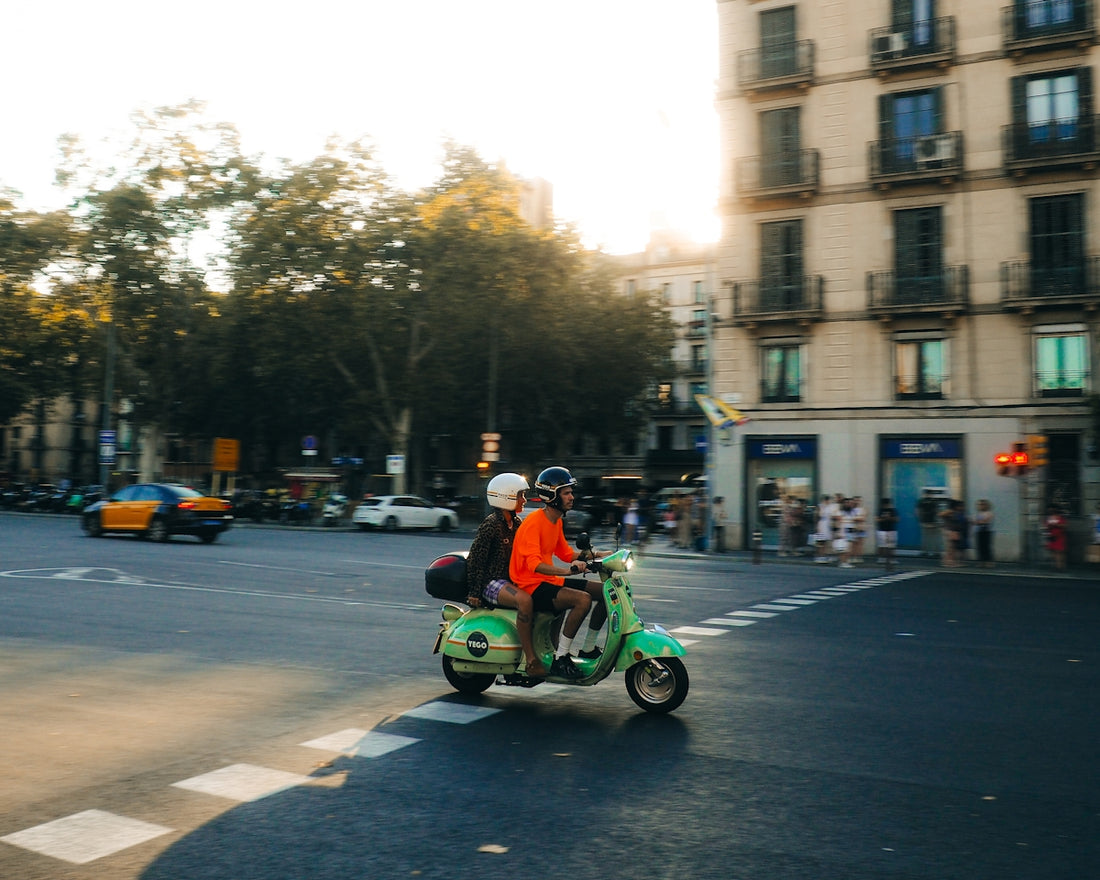 Two people ride a green scooter through a city intersection.